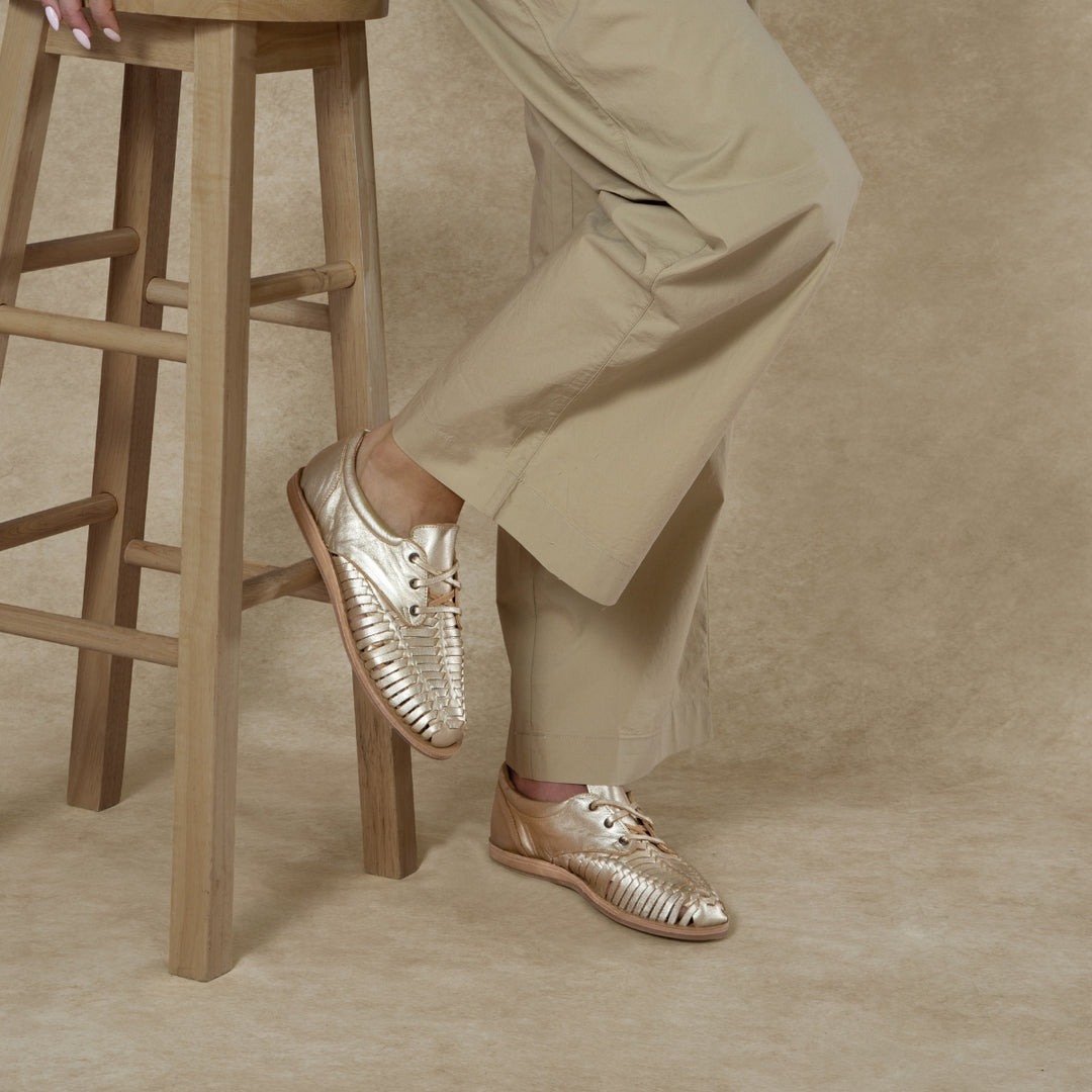 Pair of gold woven leather Huarache Oxford shoes worn with tan trousers next to a wooden stool.